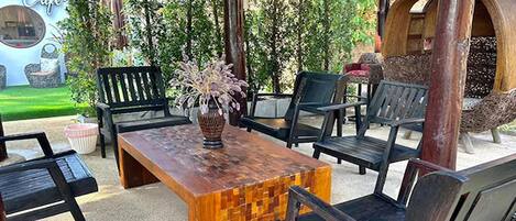 Un espacio al aire libre con mesa de madera y sillas negras, rodeado de follaje y una hamaca.