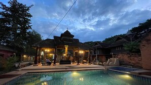 A swimming pool with tiled flooring, a covered seating area with lights, and a traditional building with a pointed roof. - Baan Tawai Lanna Resort (Hang Dong)