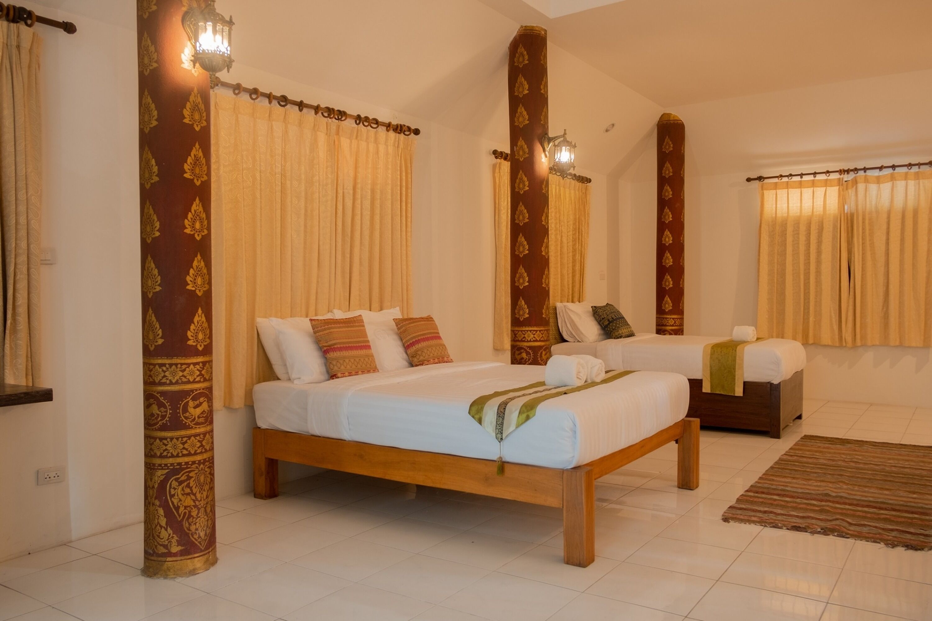 Une chambre d’hôtel avec un grand lit, un lit plus petit et un coin salon. Les fenêtres sont dotées de rideaux et il y a une lampe fixée au mur.