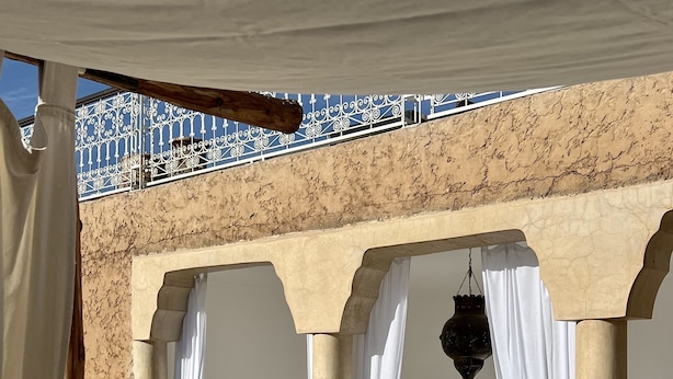 Un espacio al aire libre iluminado por el sol con cojines blancos y una mesa, enmarcado por arcos con telas colgantes.