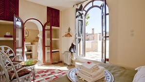 Una habitación con cama, una silla, una mesa con vasos, un radiador y vistas a través de una puerta abierta.