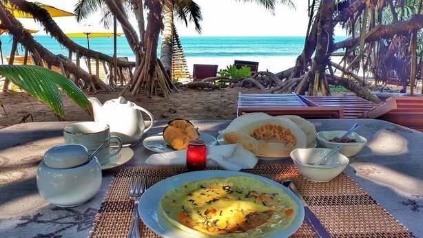 A table set with a meal, including a plate of soup, bowls, and a teapot, on a beach with palm trees and a view of the ocean. - Arcade Beach Hotel (Induruwa)