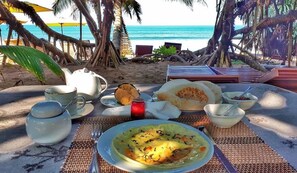 A table set with a meal, including a plate of soup, bowls, and a teapot, on a beach with palm trees and a view of the ocean. - Arcade Beach Hotel (Induruwa)