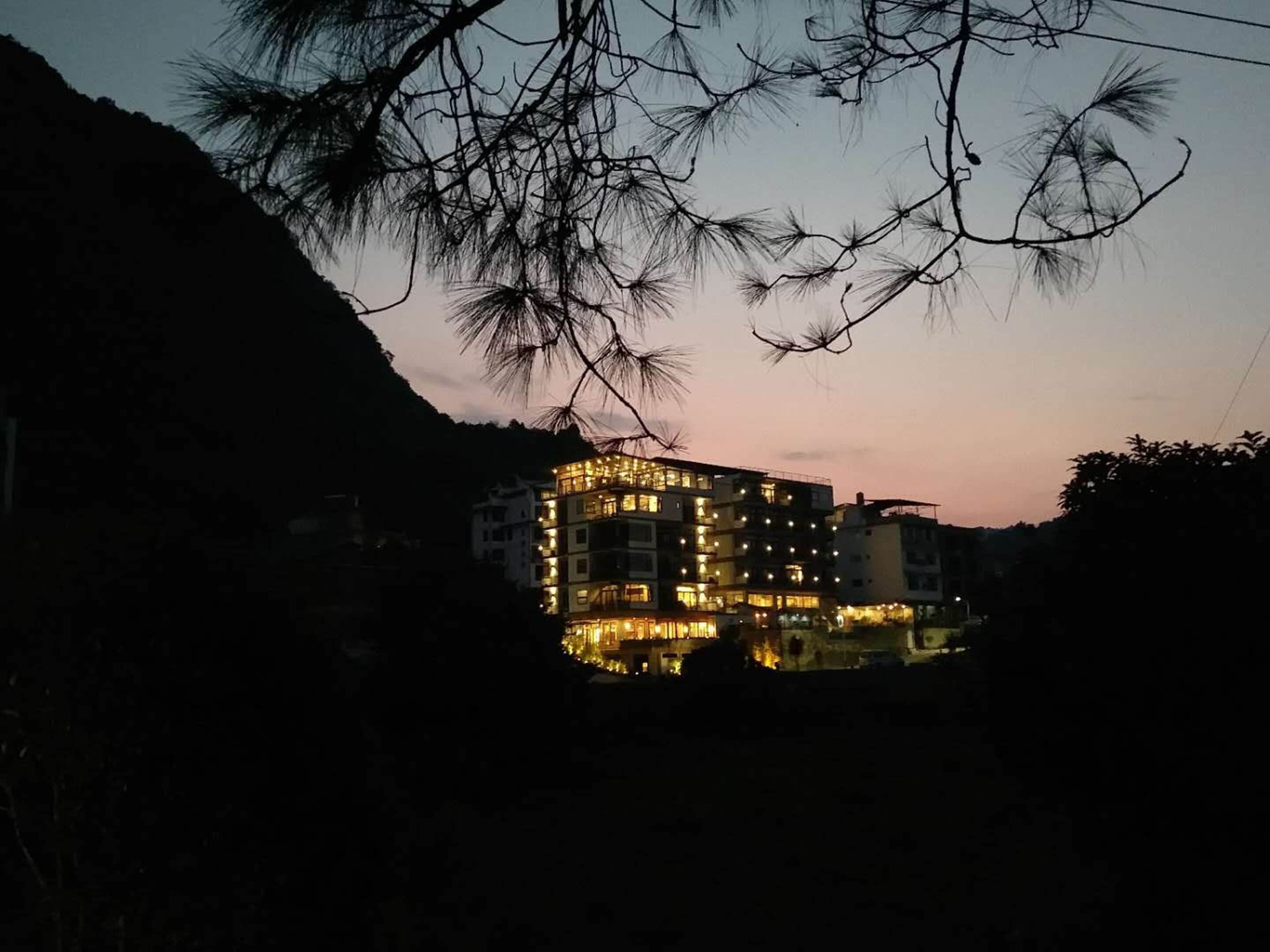 Un edificio de varios pisos con ventanas iluminadas, rodeado de árboles y colinas.