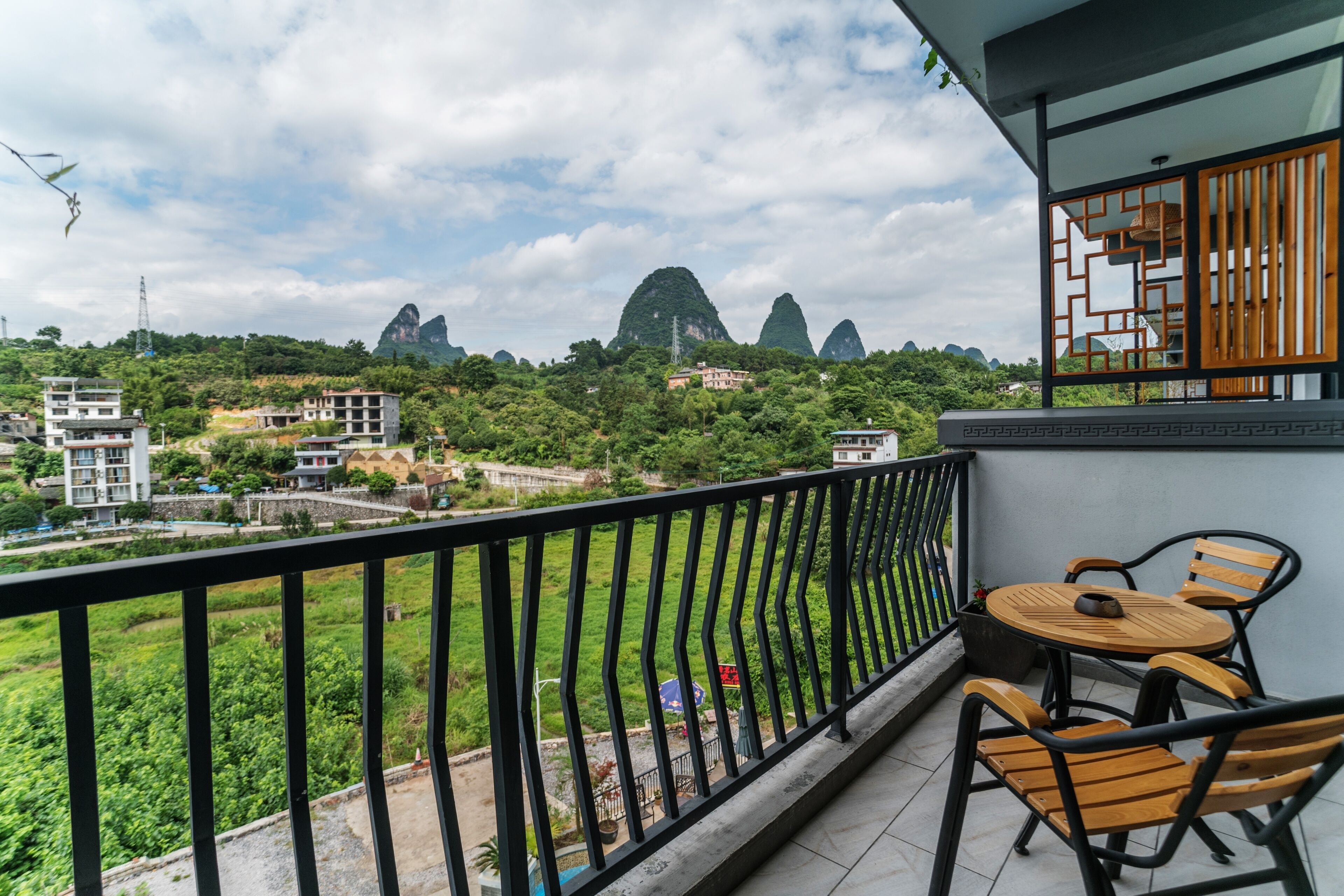 A balcony with a table and chairs, overlooking a green landscape and buildings.