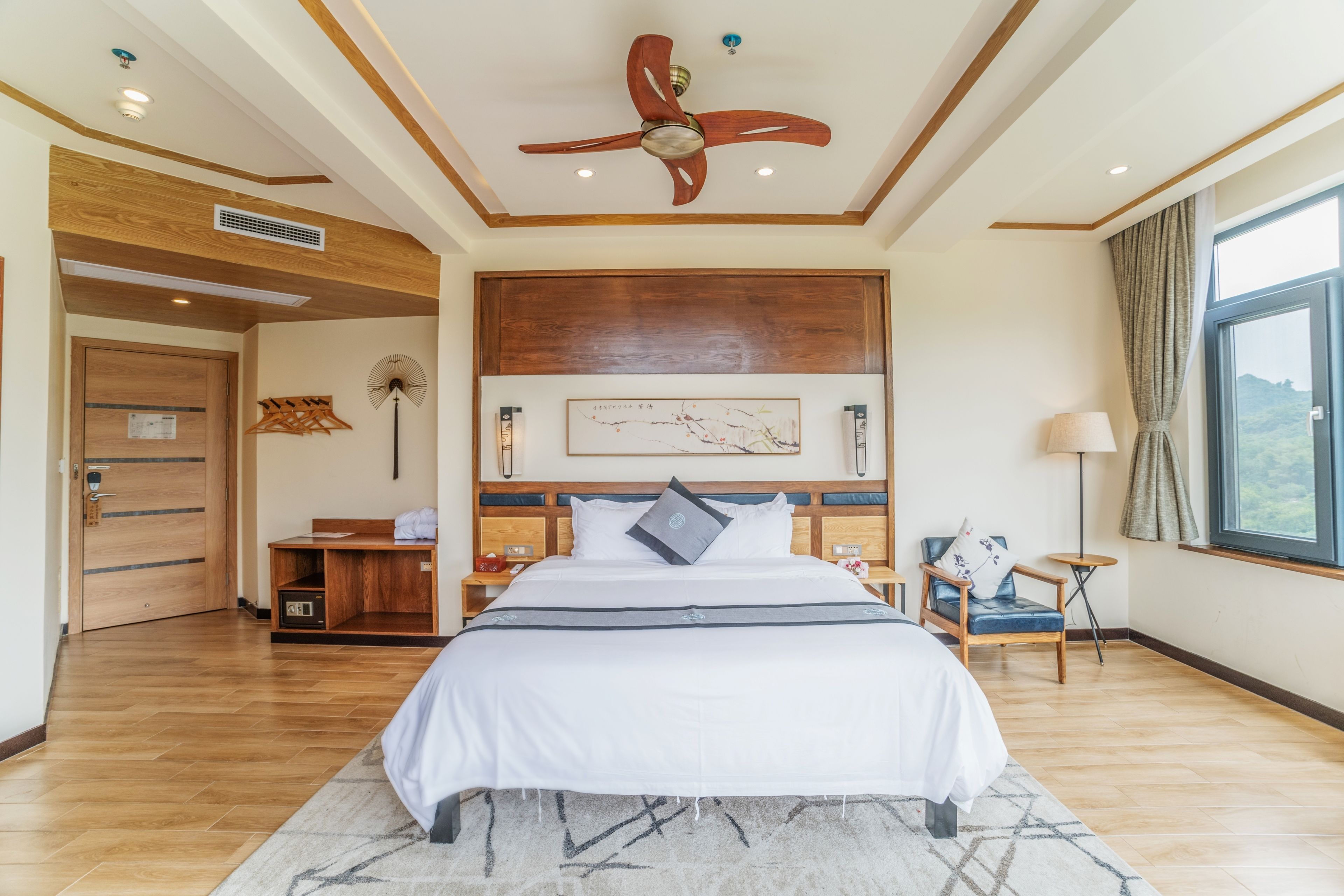 A spacious bedroom with a large bed, wooden ceiling fan, and a view of the outdoors through a window.