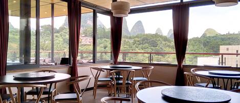 A restaurant with round tables and chairs, large windows with curtains, and a view of green hills.