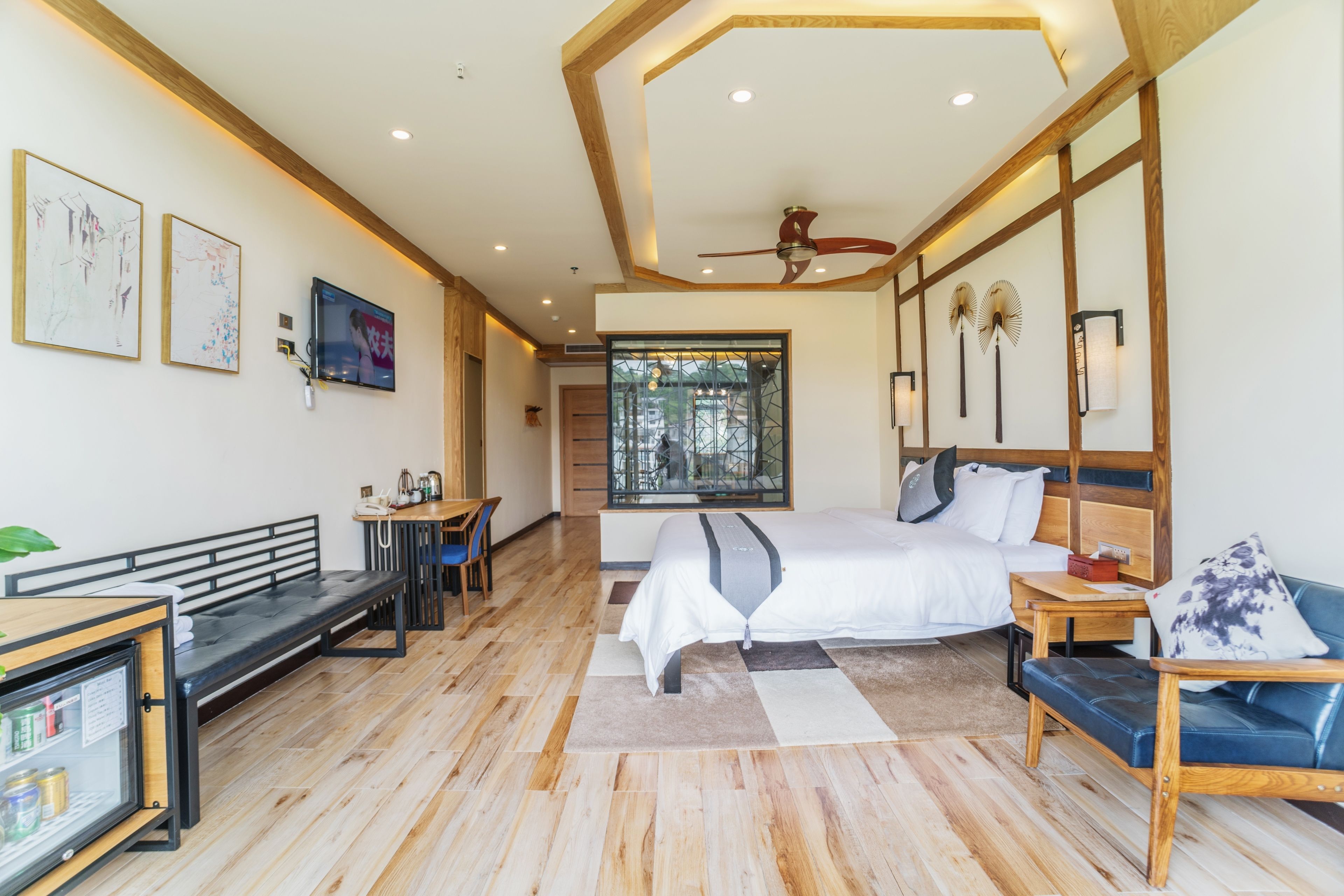 Una habitación de hotel moderna con una cama grande, un escritorio, una banca y un sofá. Hay un televisor montado en la pared, un ventilador de techo y una ventana con vistas al exterior.