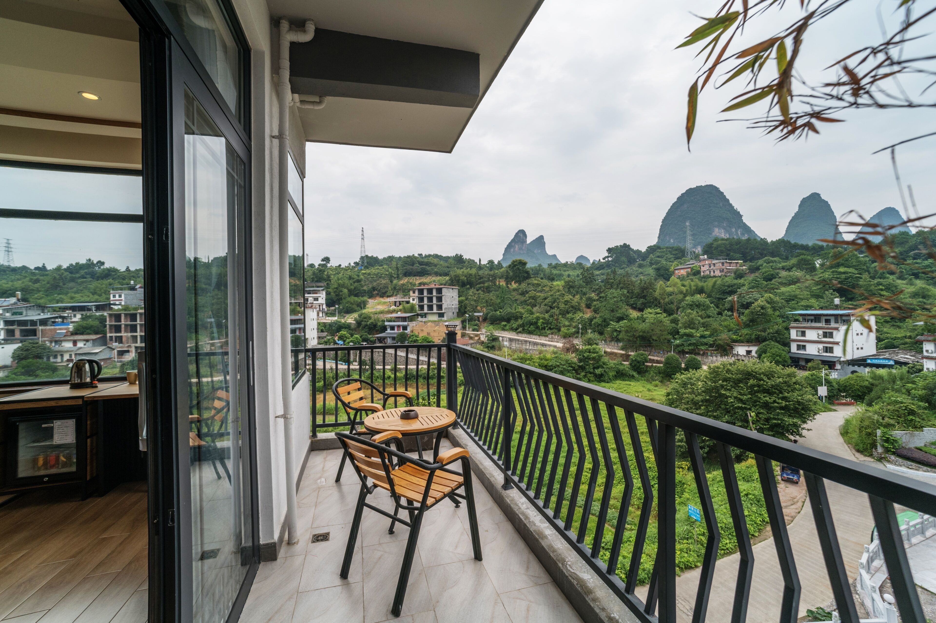 A balcony with a table and chairs, overlooking a lush green landscape with buildings and mountains in the distance.