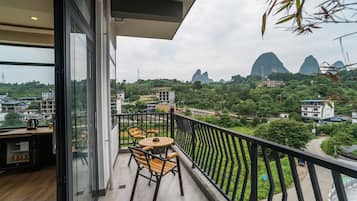 A balcony with a table and chairs, overlooking a lush green landscape with buildings and mountains in the distance.