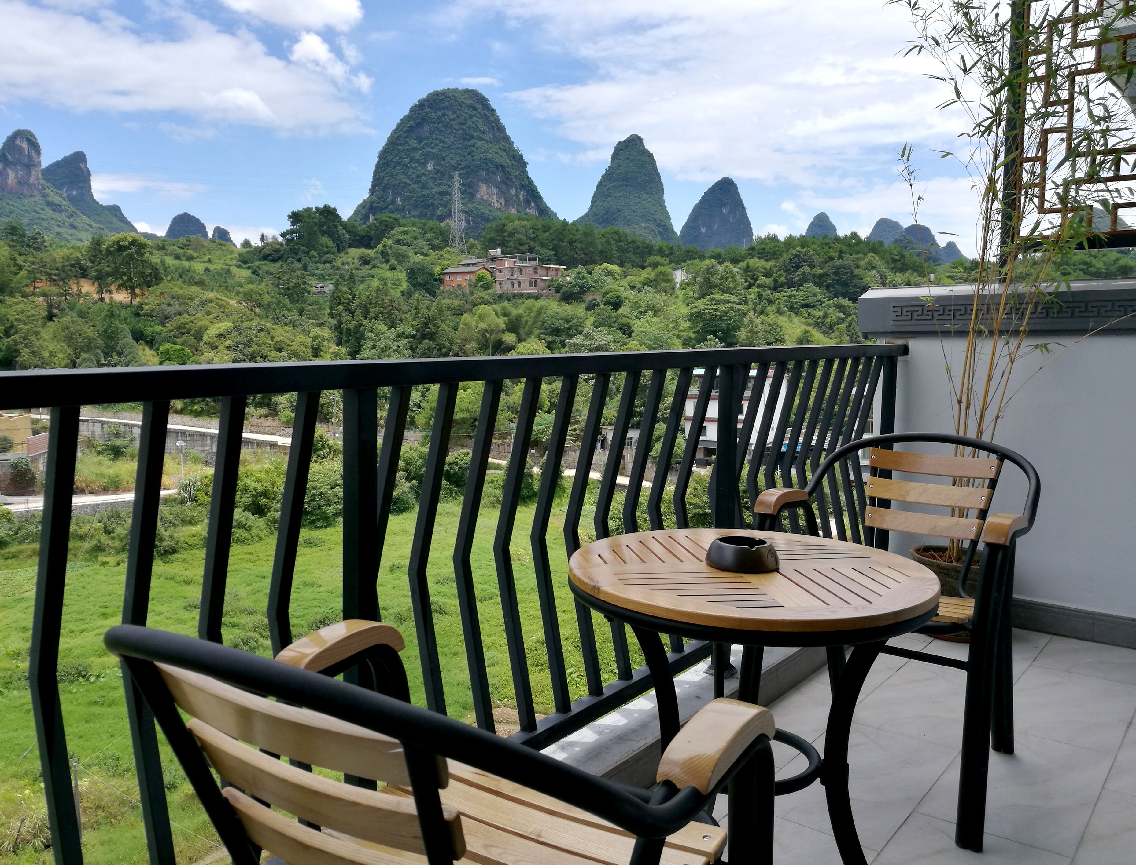 A balcony with a round table and two wooden chairs, overlooking a lush green landscape and distant mountains.