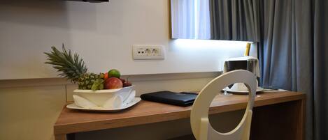 A hotel room with a wooden desk, a white chair, a fruit bowl, a laptop, a remote control, and a flat-screen TV.