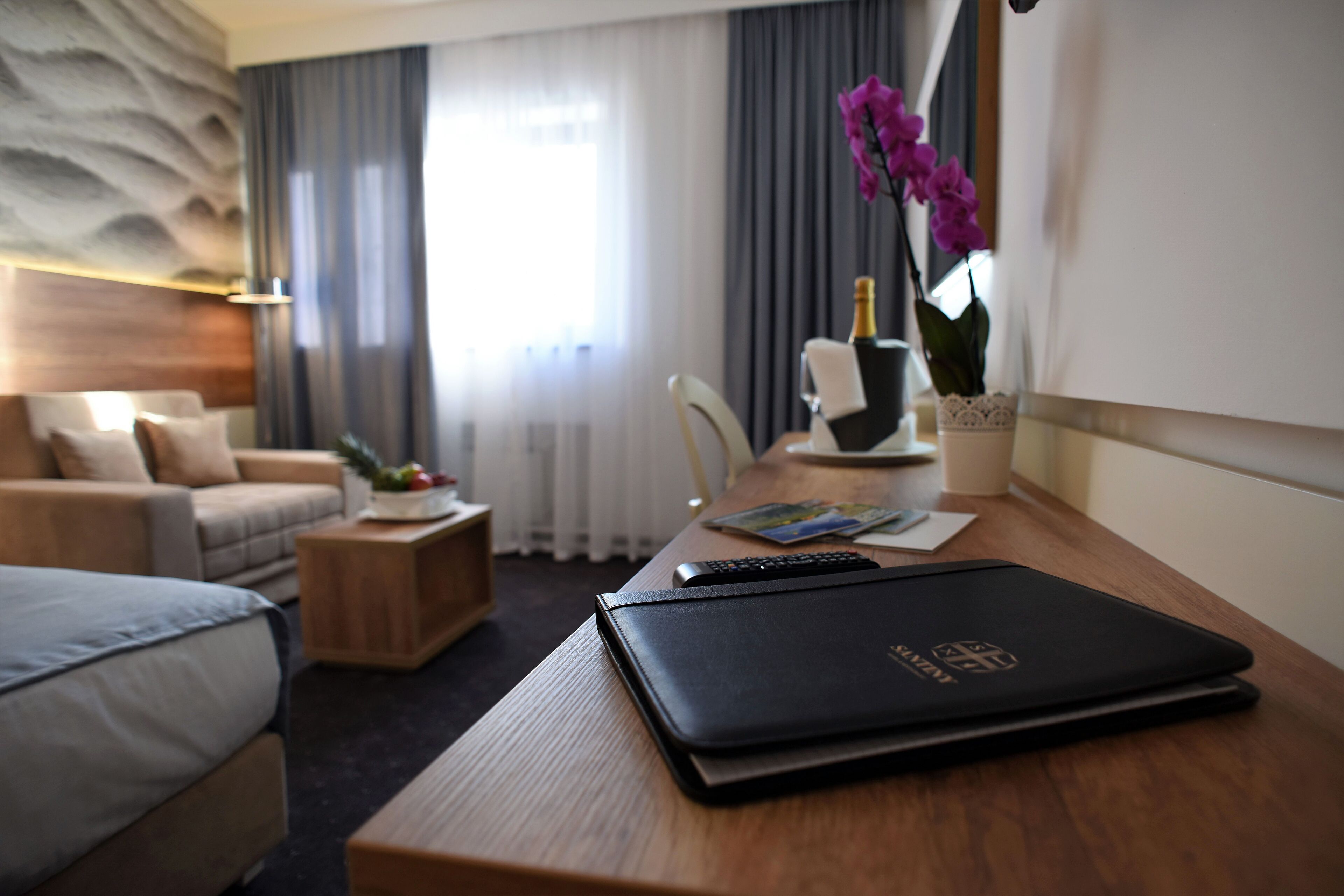 Habitación de hotel con cama, un escritorio con una computadora portátil, una silla, una mesita con un florero y una ventana con cortinas.