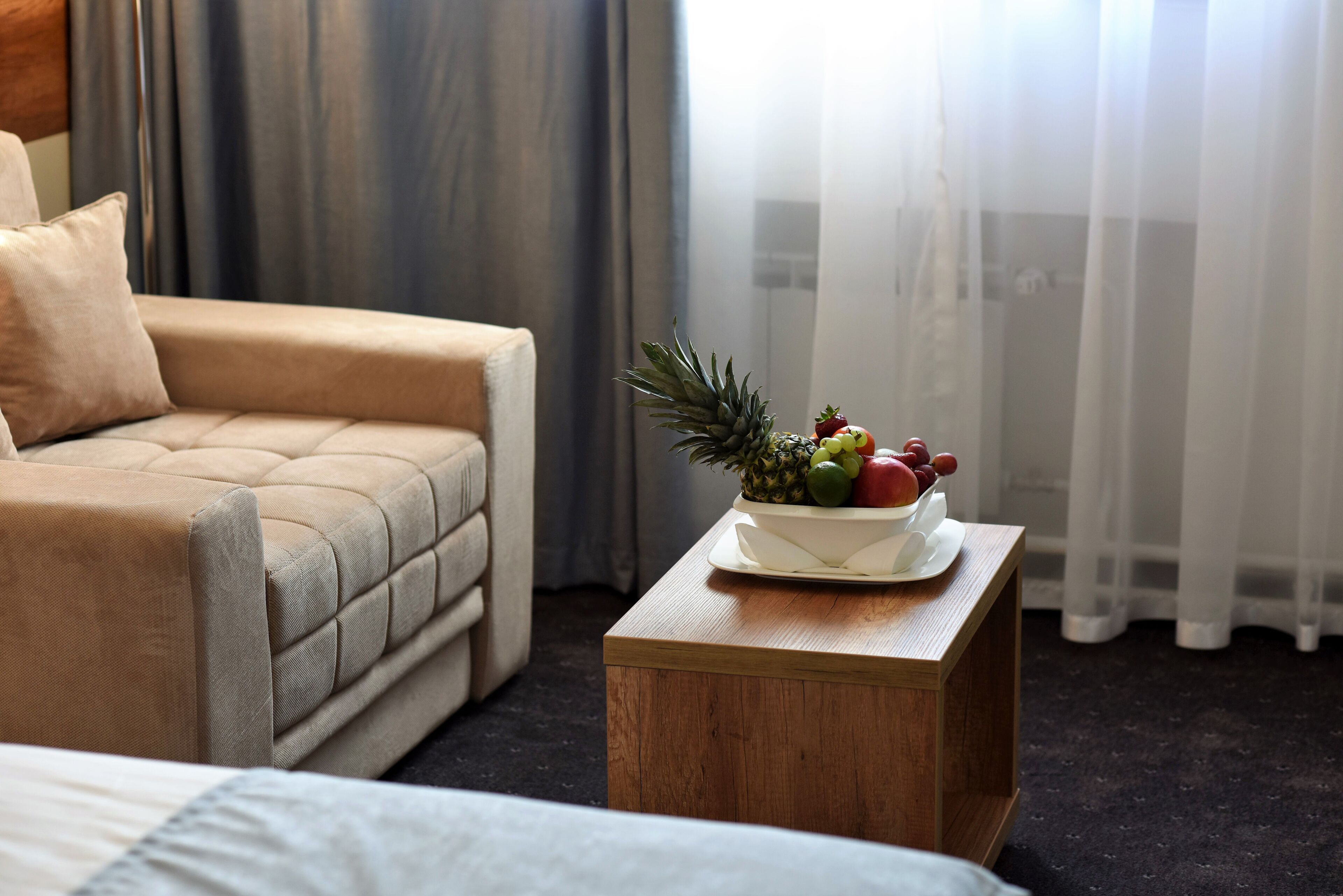 A hotel room with a bed, a sofa, a wooden nightstand with a fruit bowl, and a window with sheer curtains.