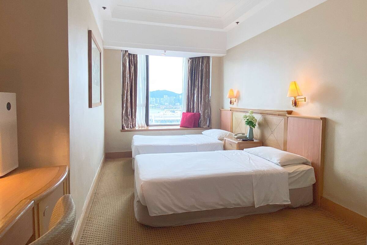 Ein Hotelzimmer mit zwei Betten, einem Schreibtisch, einem Stuhl, einem Fenster mit Vorhängen und einer Lampe.