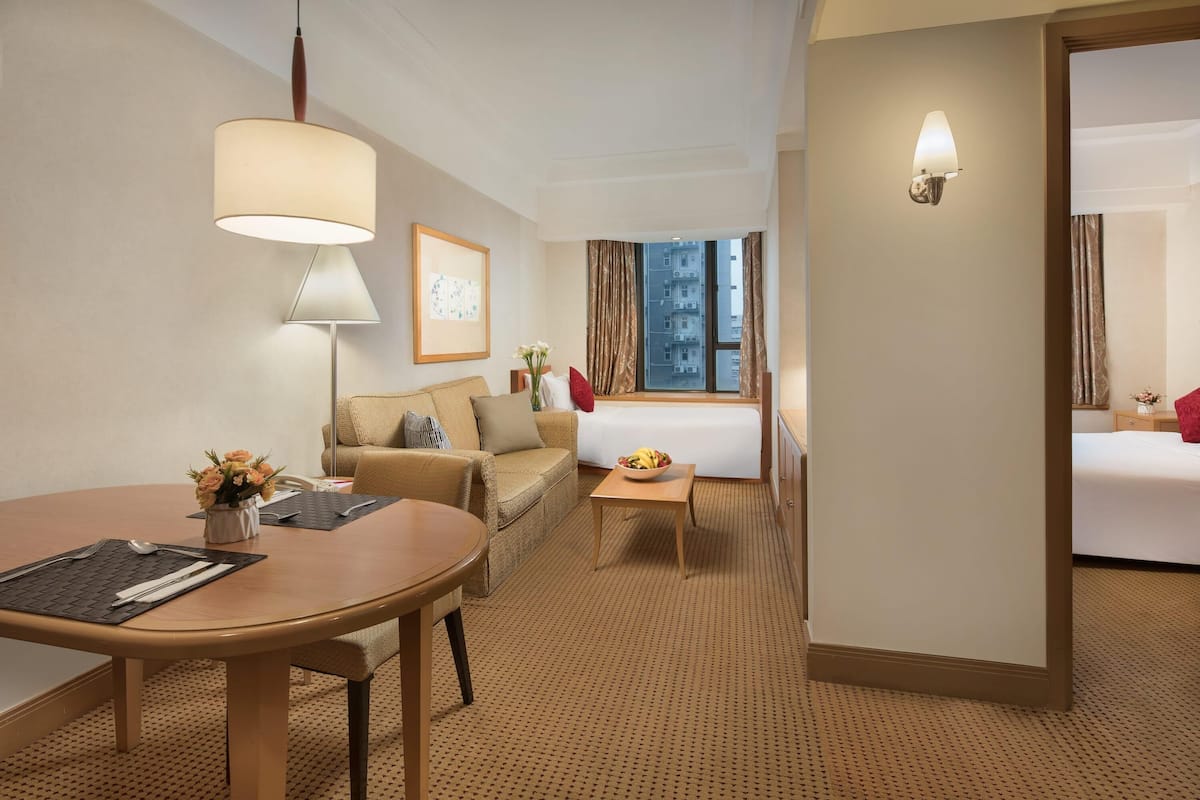 Ein Hotelzimmer mit Esstisch, einer Couch, einem kleinen Tisch mit Obstschale, einem Bett und einem Fenster mit Vorhängen.