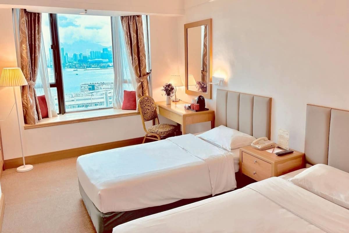 Ein Hotelzimmer mit zwei Betten, einem Schreibtisch, einem Stuhl, einer Lampe, einem Spiegel und Blick auf die Stadtlandschaft.