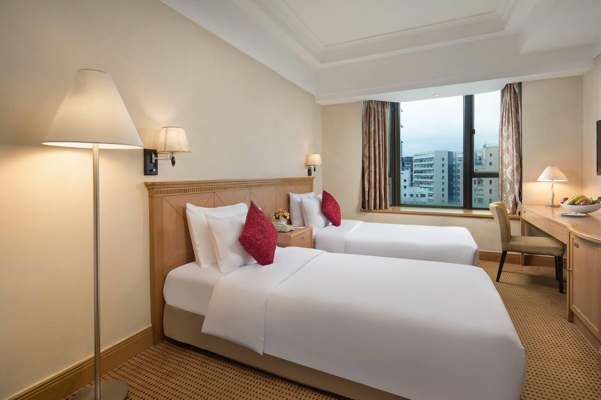 Ein Hotelzimmer mit zwei Betten, einem Schreibtisch, einem Stuhl, einer Lampe und Blick auf die Stadt.