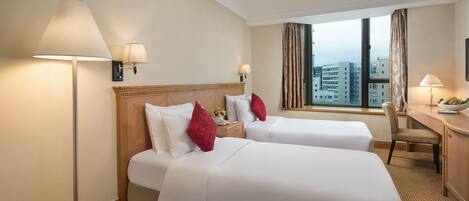 A hotel room with two beds, a desk, a chair, a lamp, and a view of the city.