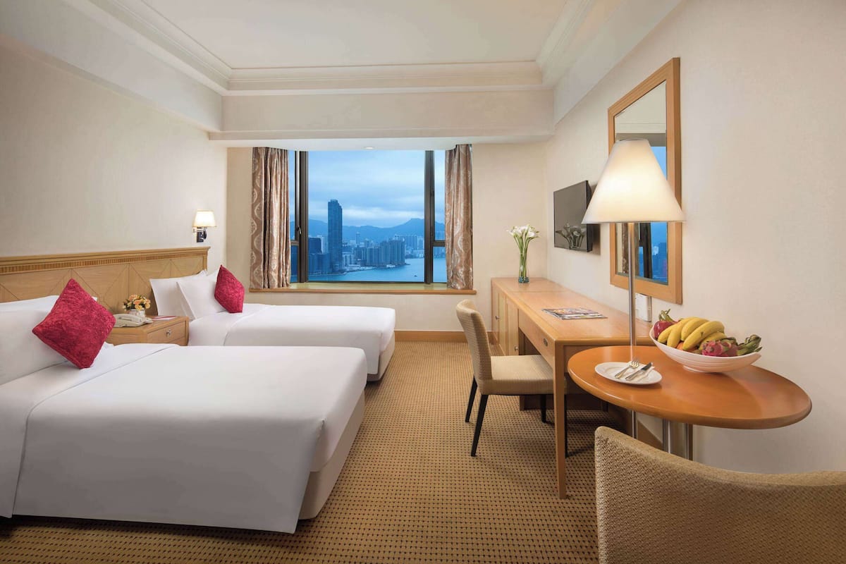 Ein Hotelzimmer mit zwei Betten, einem Schreibtisch mit Stuhl, einem Esstisch mit Obstschale, einer Lampe und einem großen Fenster mit Blick auf die Stadt.