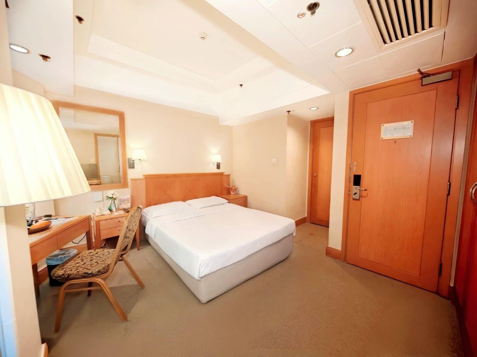 Une chambre d’hôtel avec un lit simple, un bureau avec une chaise, une lampe et une armoire en bois.