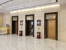 Vestíbulo con piso de mármol, mostrador de recepción y tres ascensores con letreros de First Cafe.