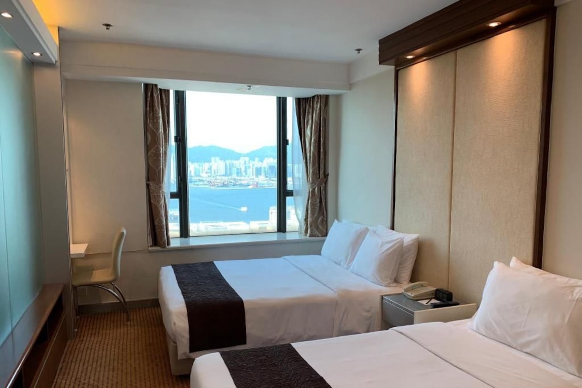 Habitación de hotel con dos camas, un escritorio, una silla, un ventanal con vista a la ciudad y un teléfono junto a la cama.
