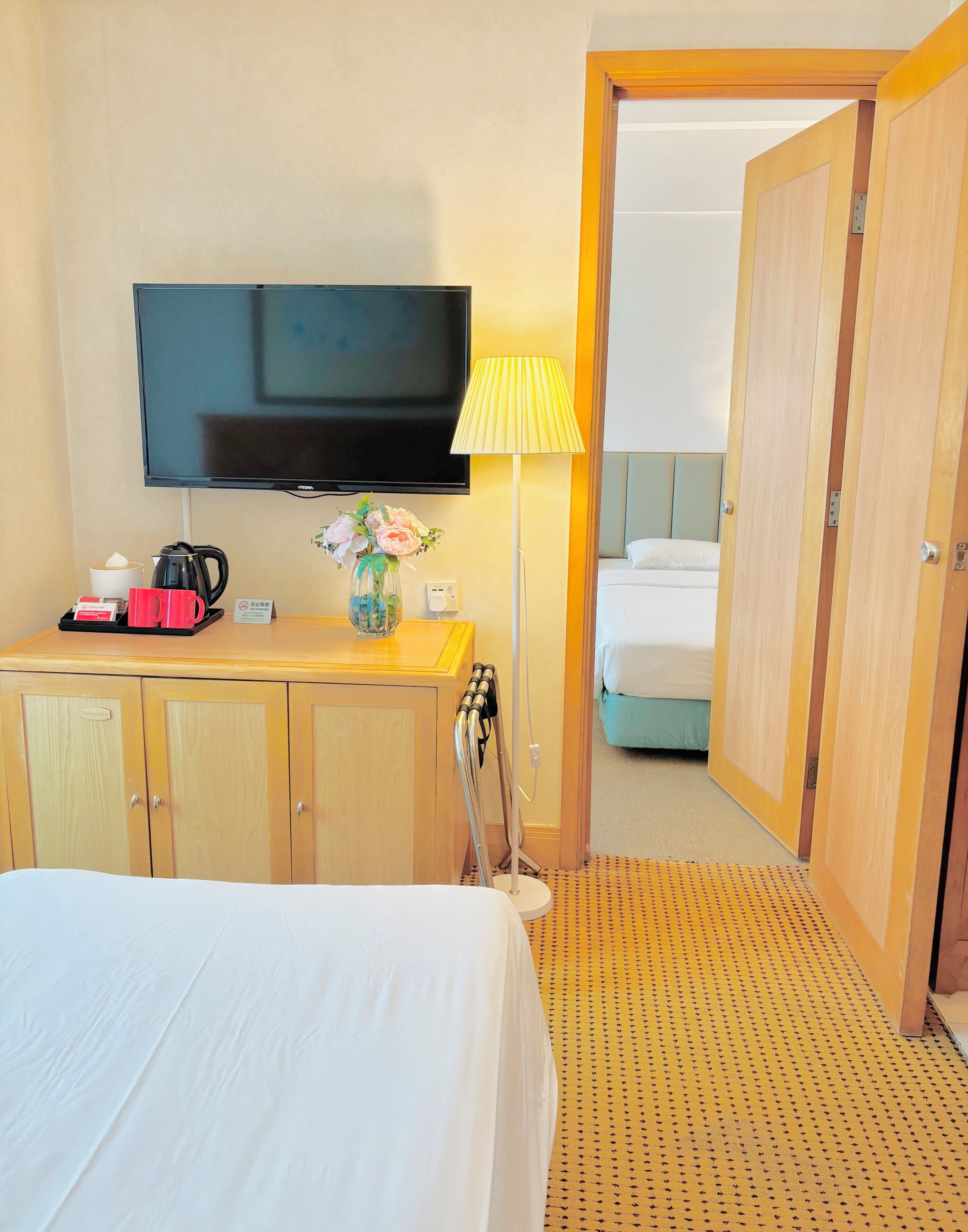 A hotel room with a bed, a TV, a desk with a coffee maker, a lamp, and a wardrobe.