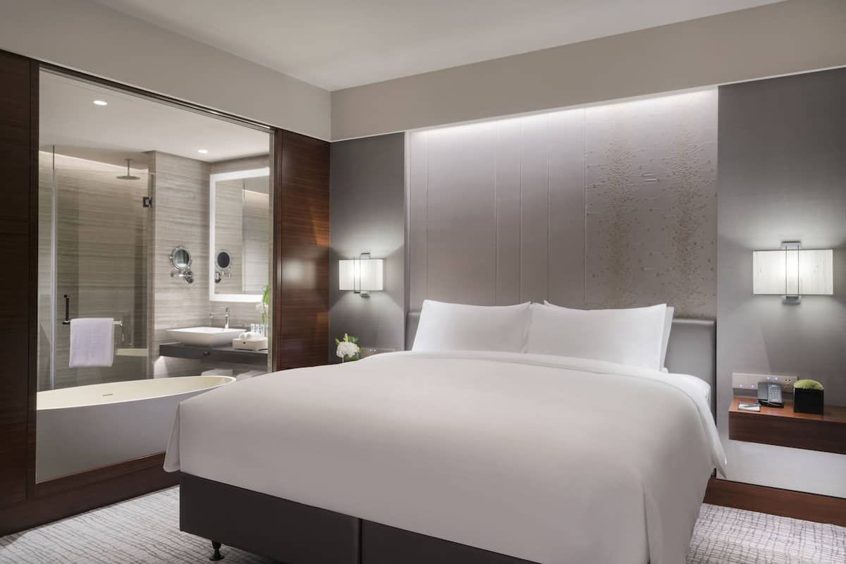 A modern hotel room with a large bed, a bathroom with a bathtub, a mirror, and two bedside tables with lamps.