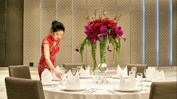 Una mesa de comedor elegantemente dispuesta con un centro de flores, una mujer con vestido rojo acomodando la vajilla, y un interior moderno con paredes decoradas.