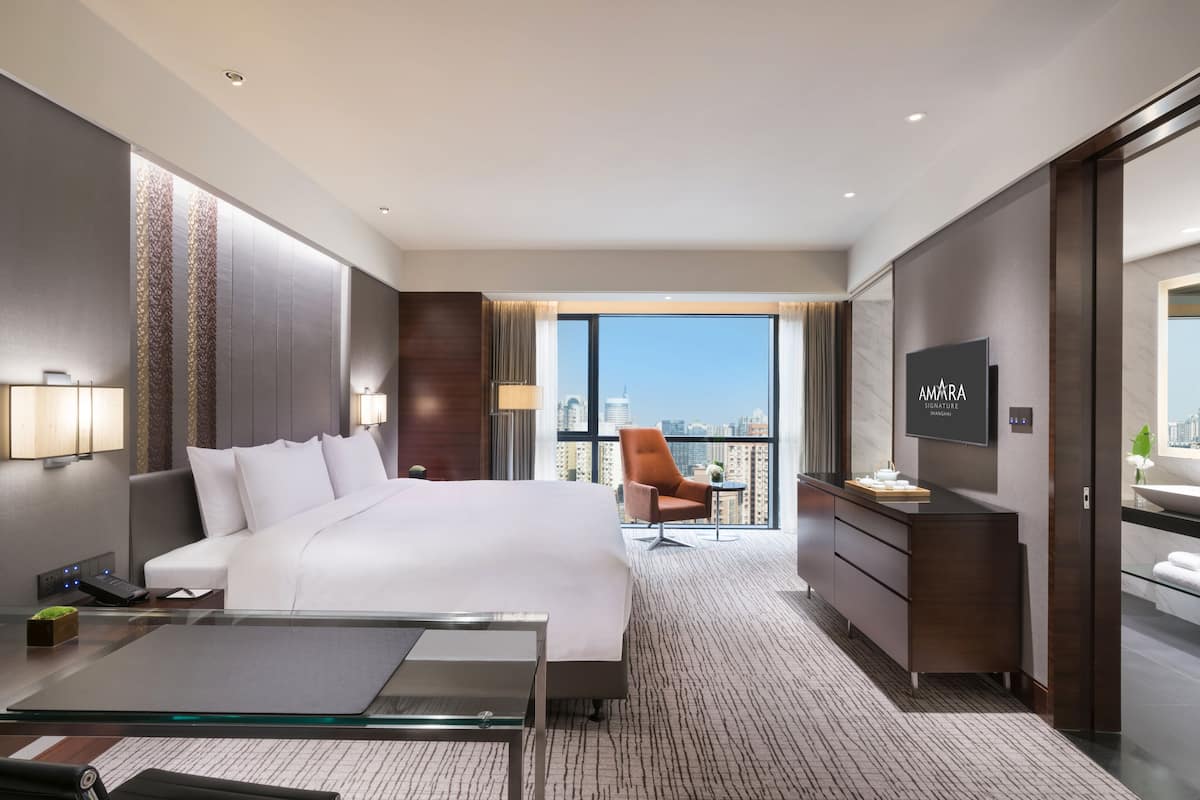 A modern hotel room with a large bed, a desk, a chair, a TV, and a balcony with a city view.