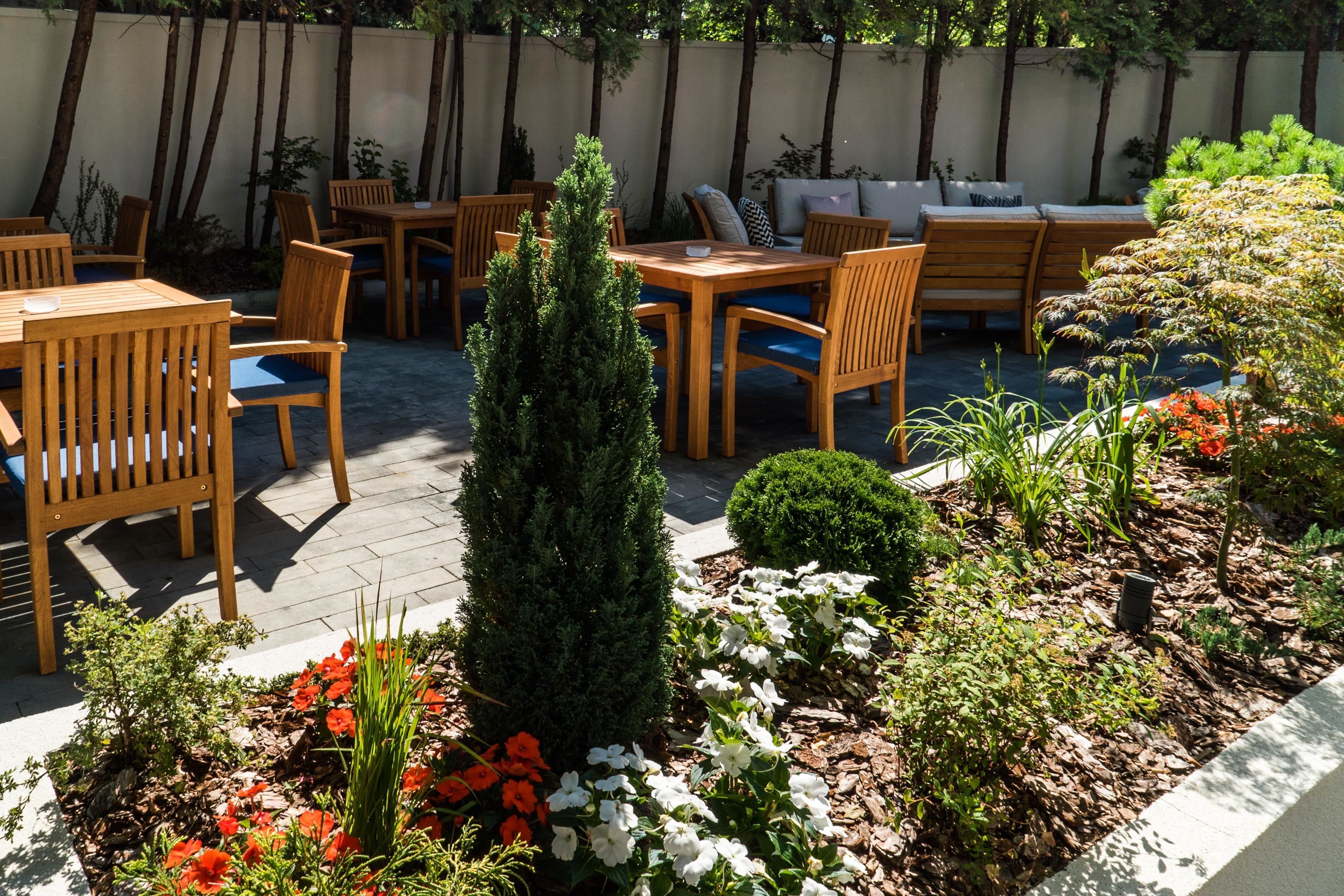 An outdoor patio area with wooden furniture, including tables and chairs, surrounded by a variety of plants and flowers.