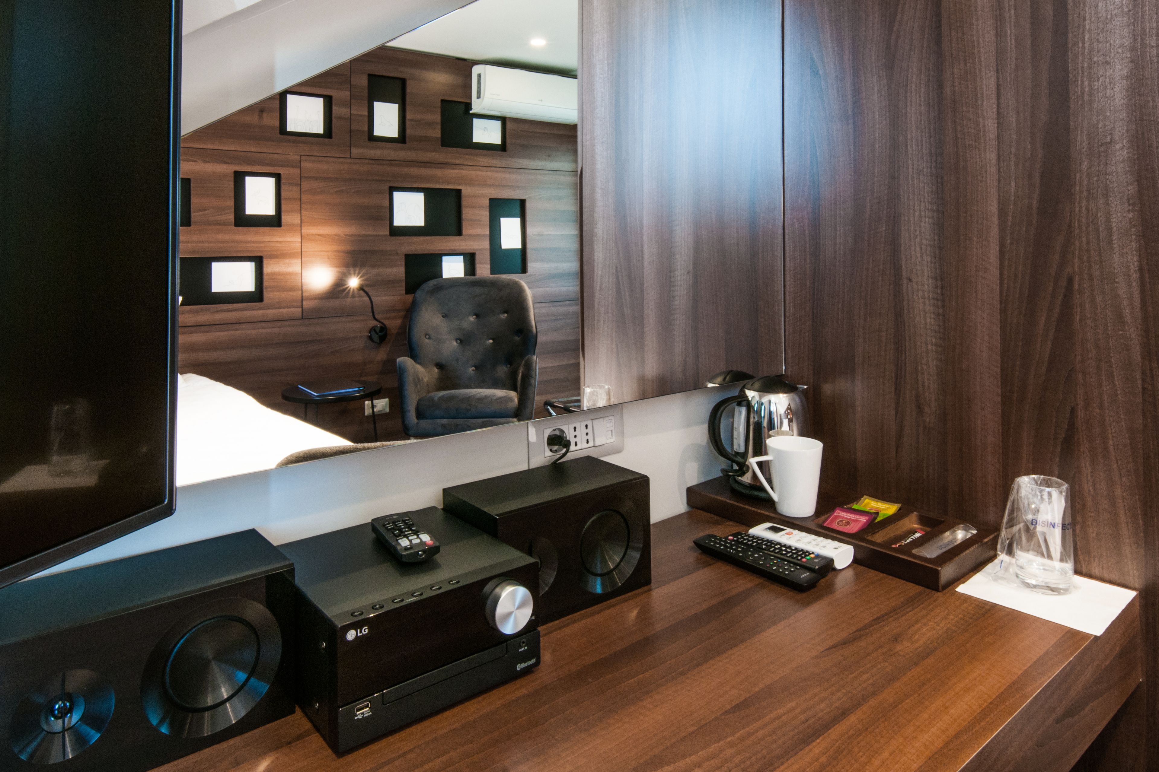 A modern hotel room with a wooden wall, a flat-screen TV, a sound system, a coffee machine, and a glass of water on the counter.