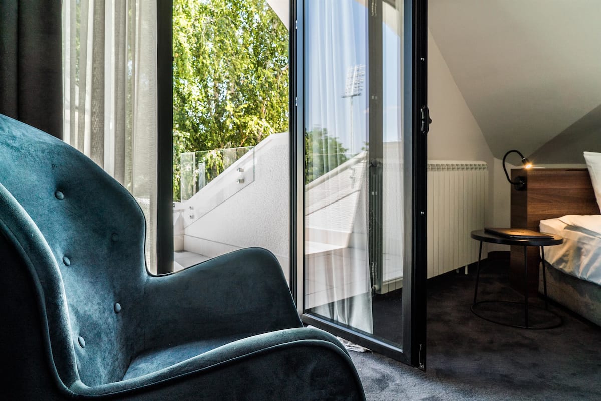 Une chambre moderne dotée d’une grande porte-fenêtre coulissante donnant sur un balcon, d’un fauteuil confortable, d’une petite table ronde et d’un lit soigneusement fait.