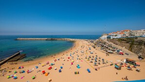 A beach with numerous colorful umbrellas and sun loungers, a rocky breakwater, and buildings with red roofs along the coastline.