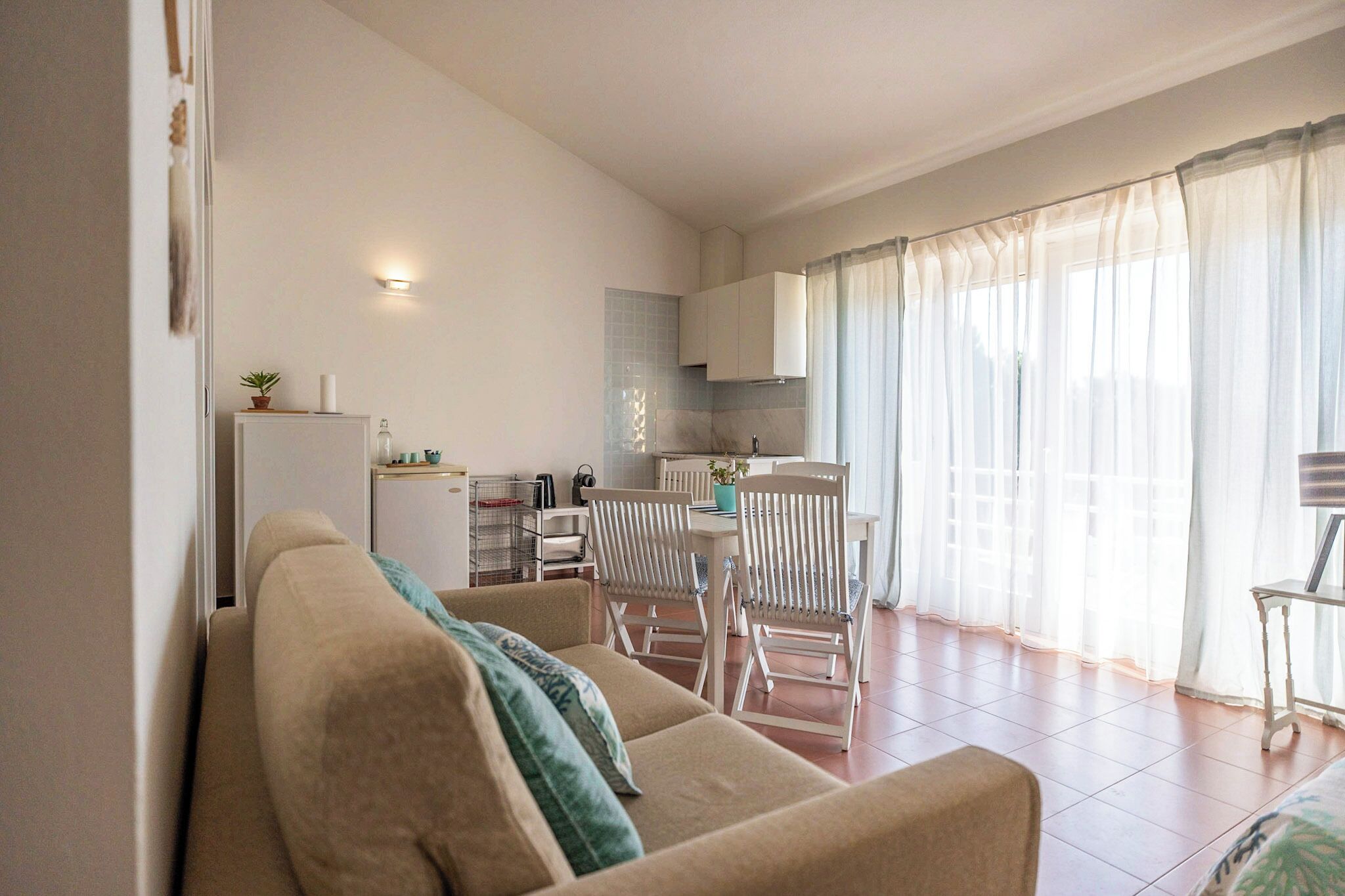 Uma sala de estar com sofá, cadeiras, uma mesa de jantar e uma área de cozinha ao fundo.