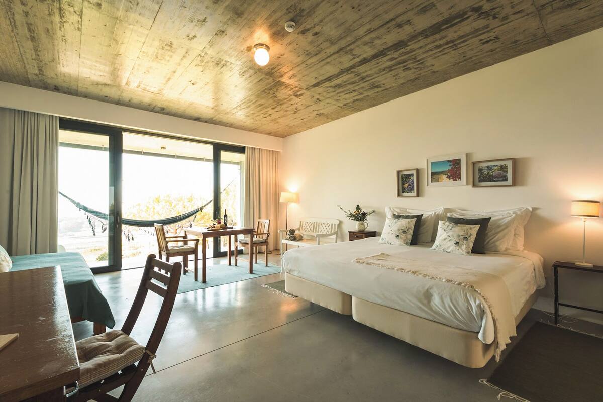 A spacious bedroom with a large bed, a dining area with a table and chairs, and a hammock by the window.