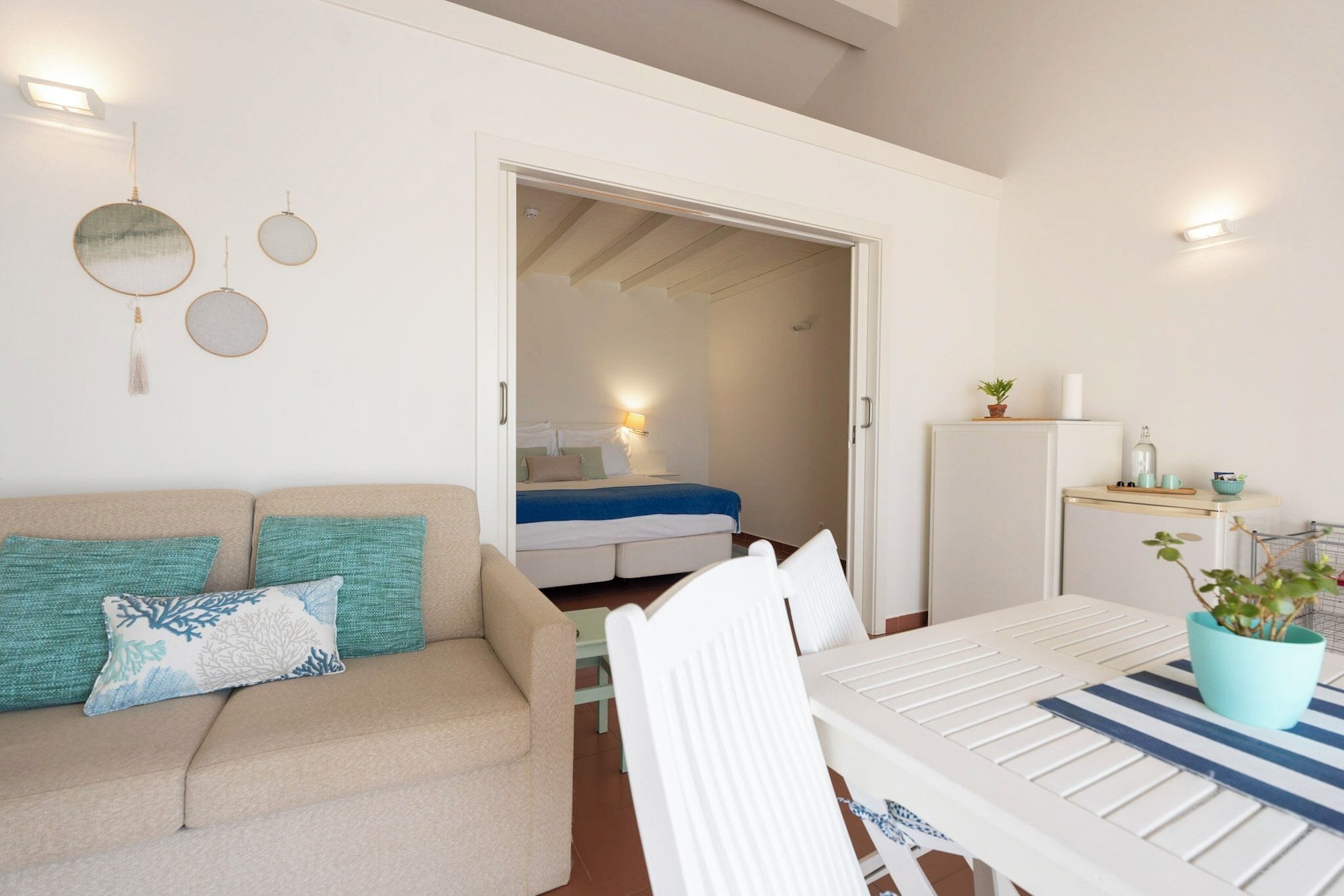 Una habitación moderna y luminosa con cama, sofá con cojines, mesa de comedor y pequeña cocina.