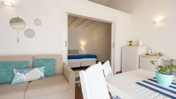 Una habitación moderna y luminosa con cama, sofá con cojines, mesa de comedor y pequeña cocina.
