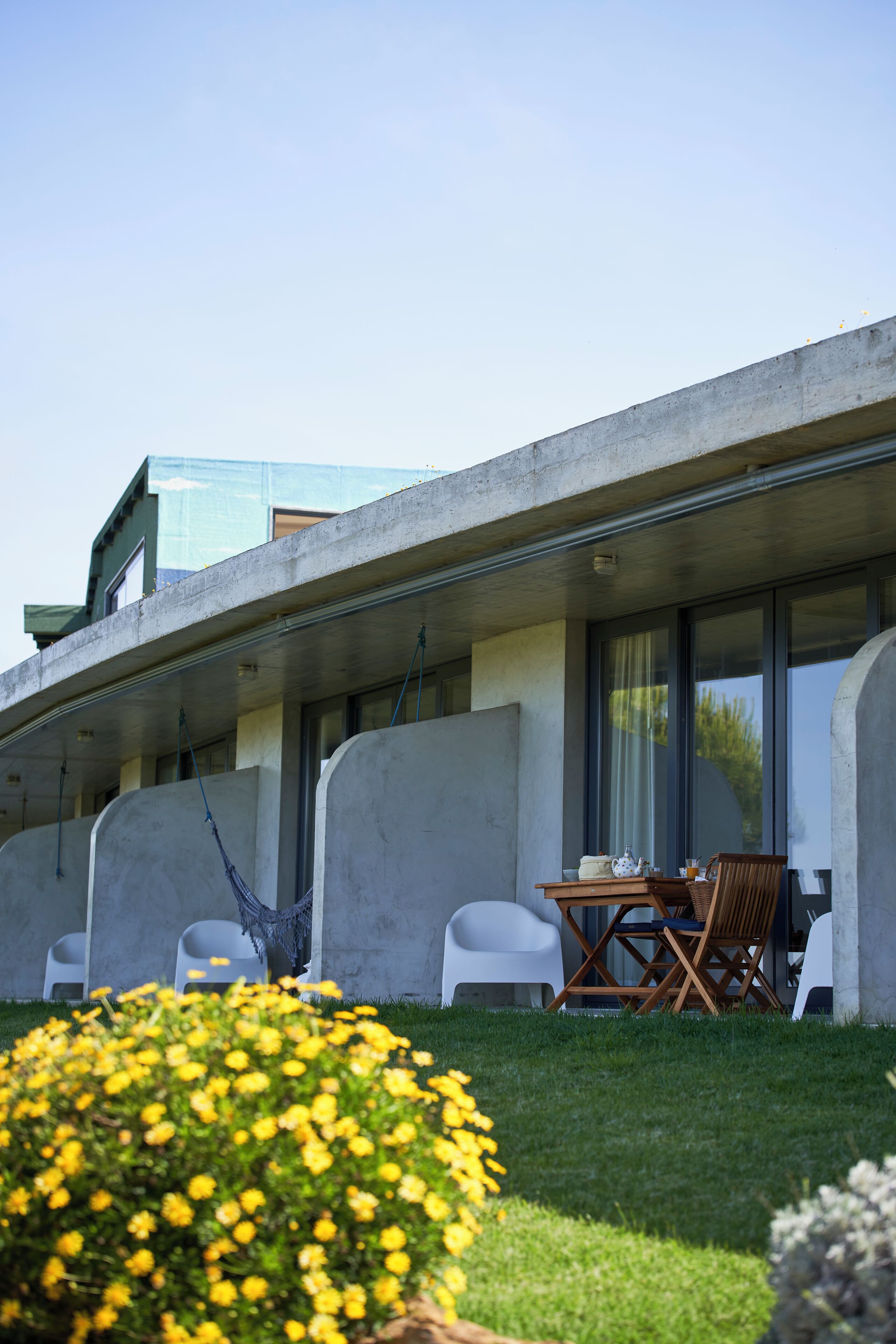 A modern building with a flat roof, large windows, and a balcony. There is a table with chairs and a hammock on the ground.