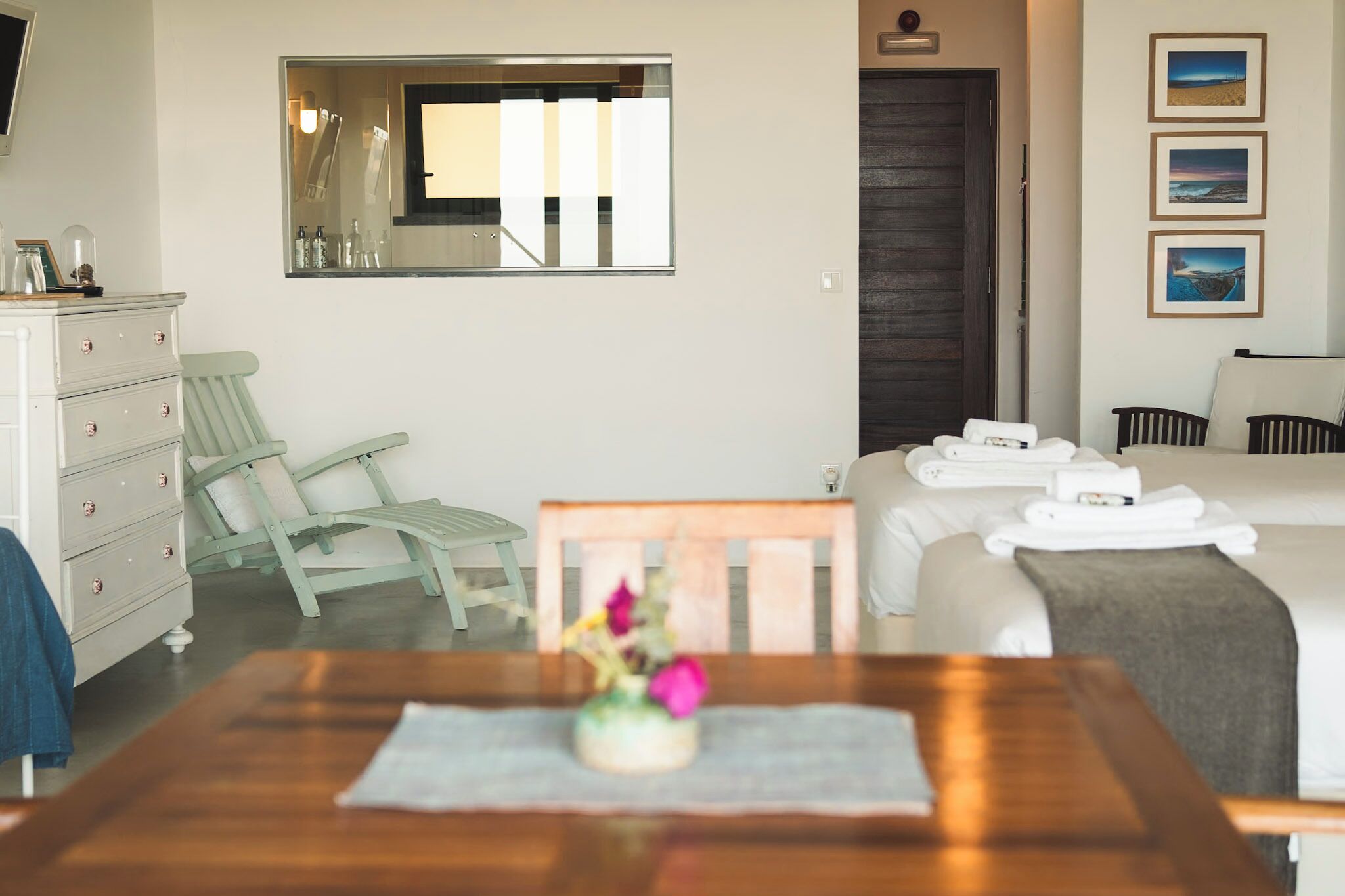 Habitación de hotel con cama, cómoda de madera, silla, mesa con un jarrón de flores, espejo y cuadros en la pared.