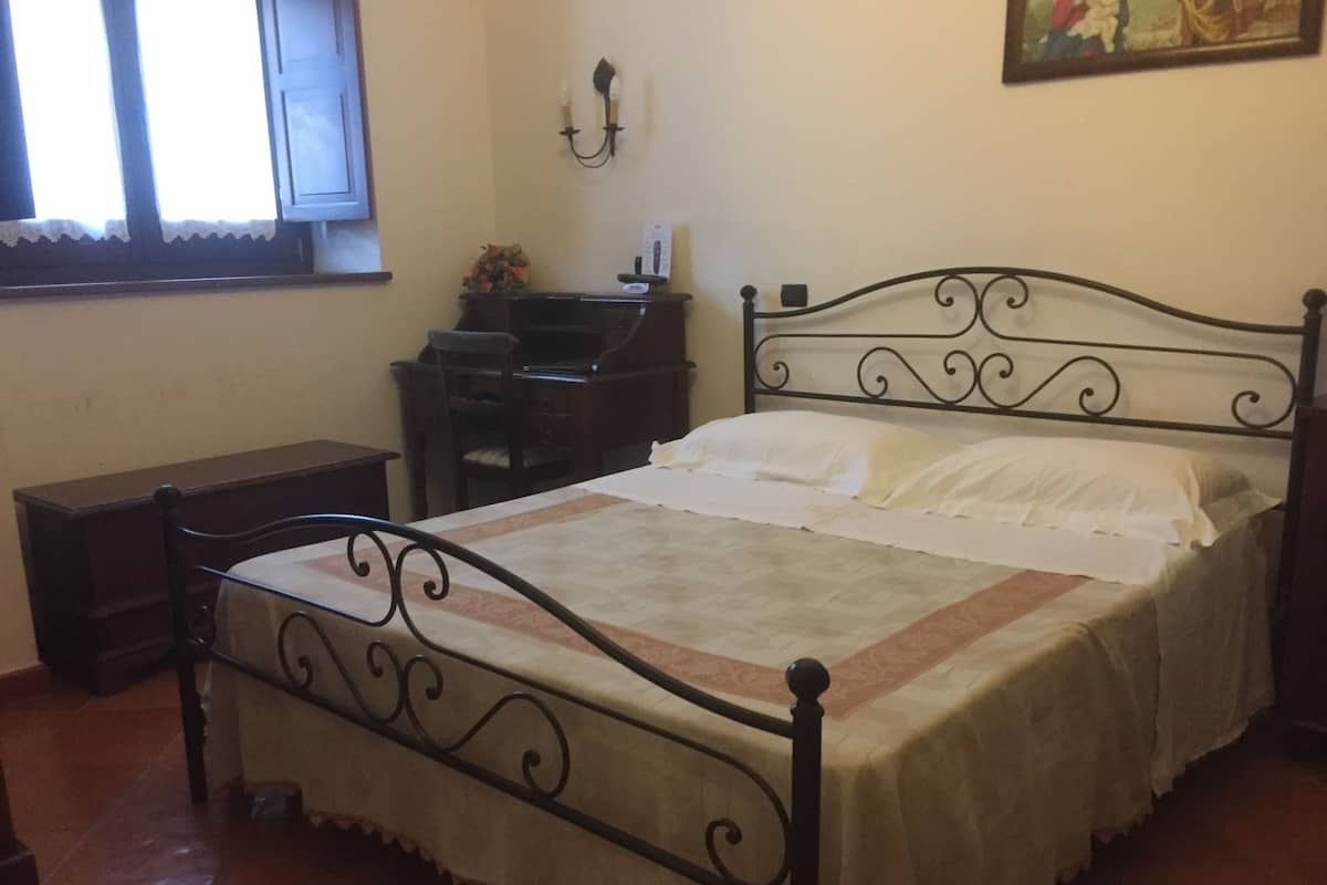 Een slaapkamer met een metalen bedframe, een houten nachtkastje, een bureau en een stoel.