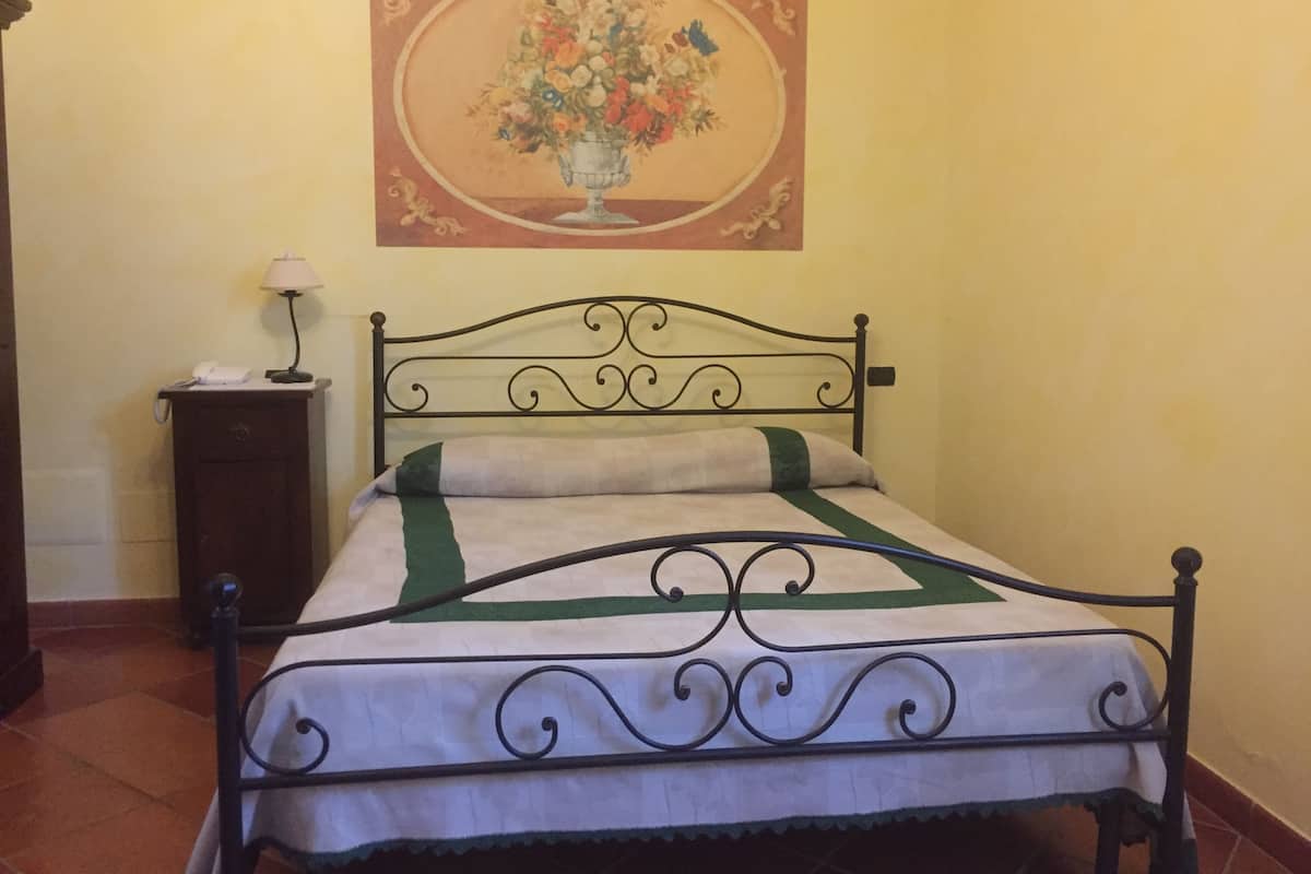Een bed met een metalen frame, een nachtkastje met een lamp en een bloemschilderij aan de muur.