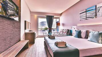 A modern hotel room with a large bed, a desk, and a balcony with a view of the mountains.