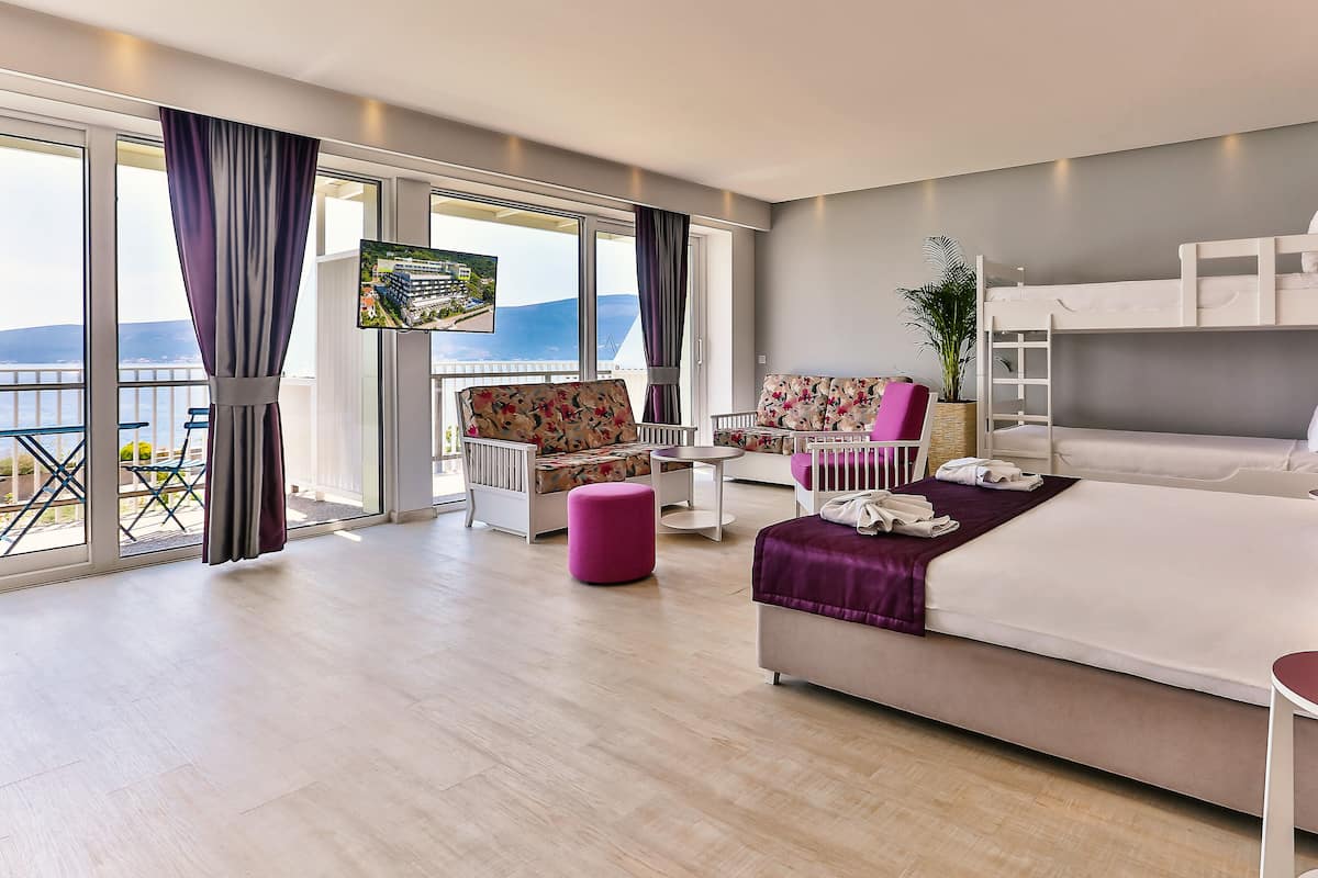 Habitación de hotel moderna con una cama grande, un balcón con televisor, zona de estar con una silla y una mesa, y vista al mar.