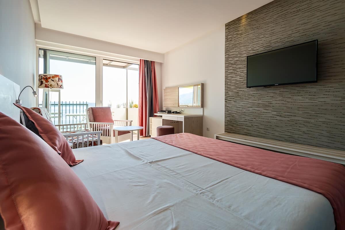 Habitación de hotel con una cama grande, un escritorio, una silla, un televisor en la pared y un balcón con vistas.