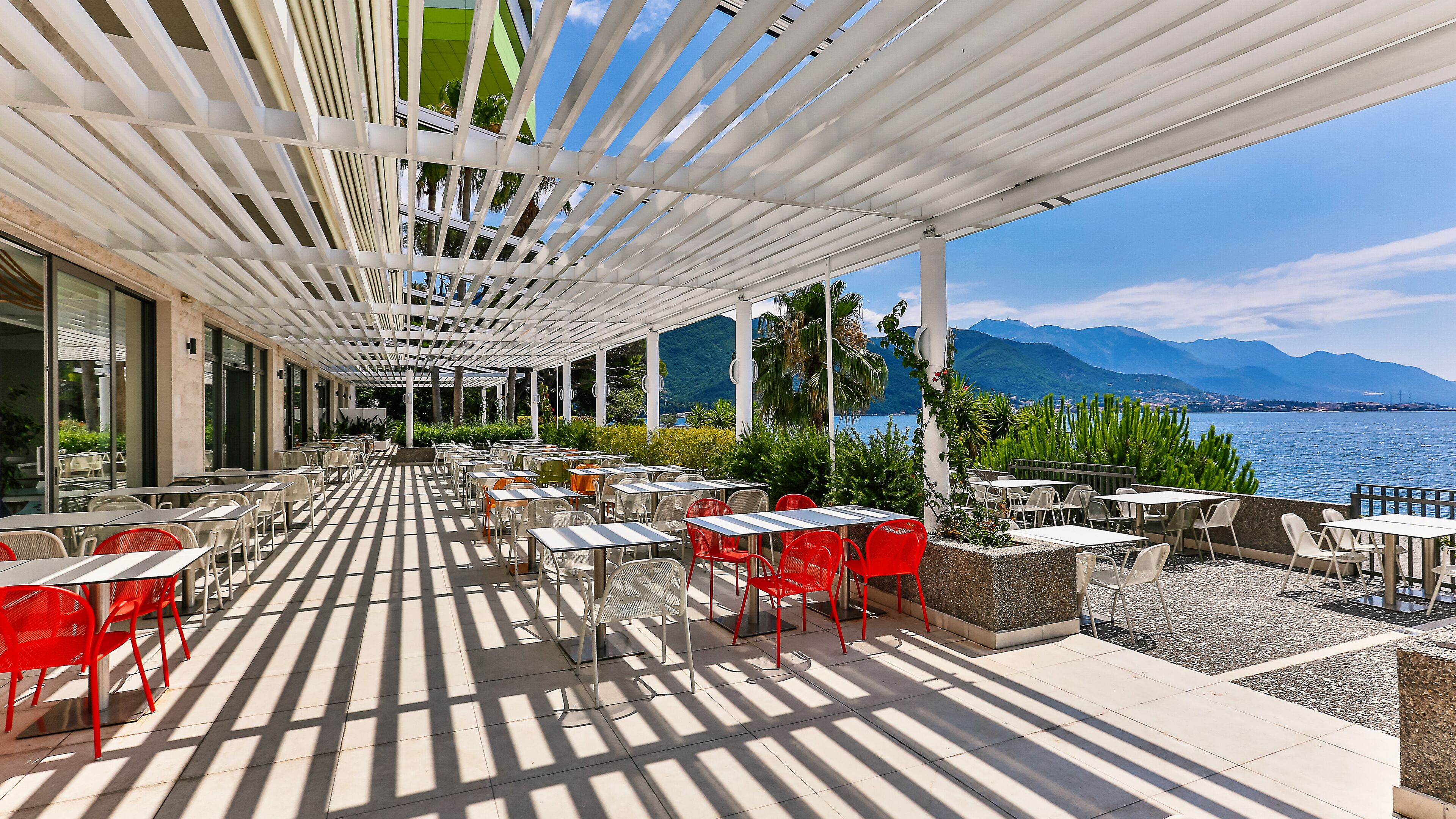 Un caffè all'aperto con tavoli e sedie, un pergolato bianco e vista su un lago e montagne.