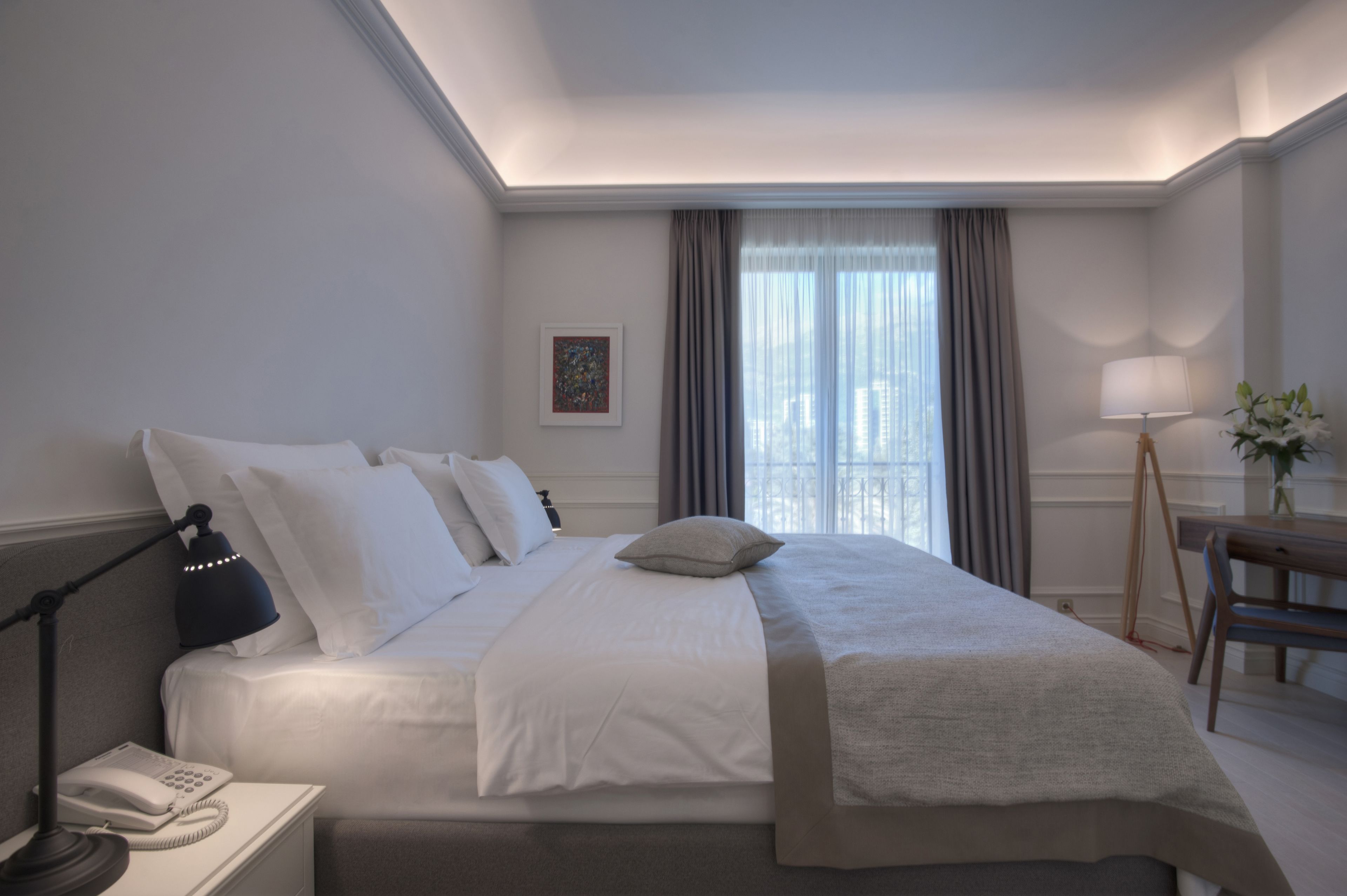 Ett hotellrum med en väl bäddad säng, en sänglampa, en telefon, ett skrivbord med en lampa och ett fönster med gardiner.