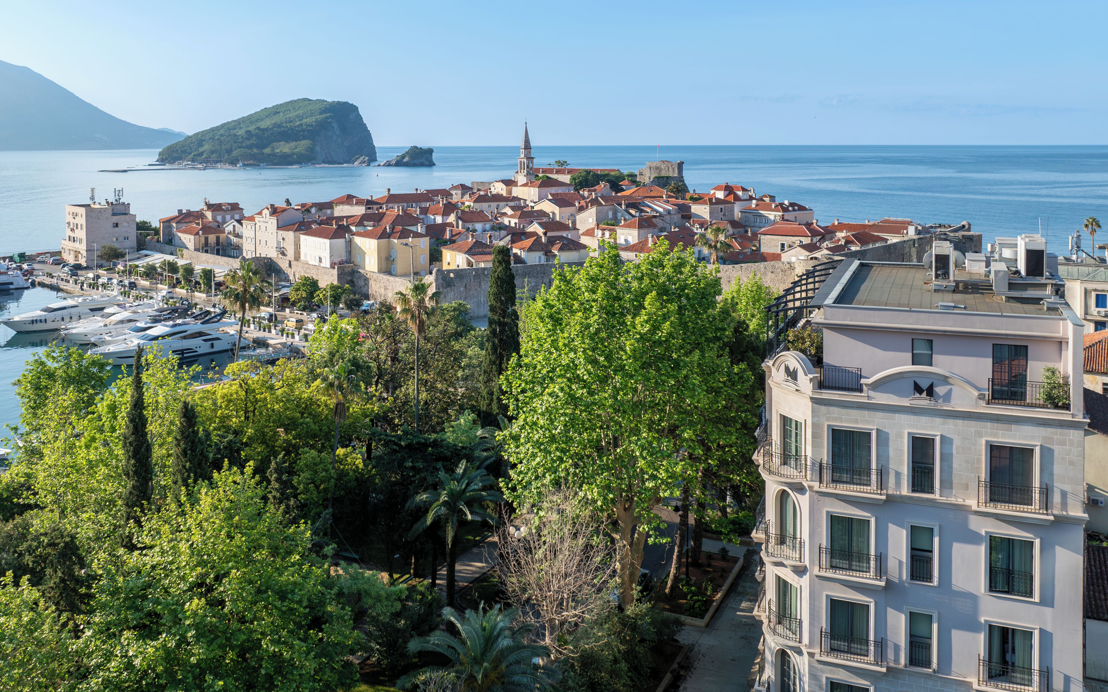 Une ville côtière dotée de bâtiments historiques, d’un port de plaisance et d’une colline imposante en arrière-plan.