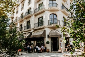 Un área de café al aire libre con mesas y sillas, rodeada por un edificio con balcones y ventanas amplias.